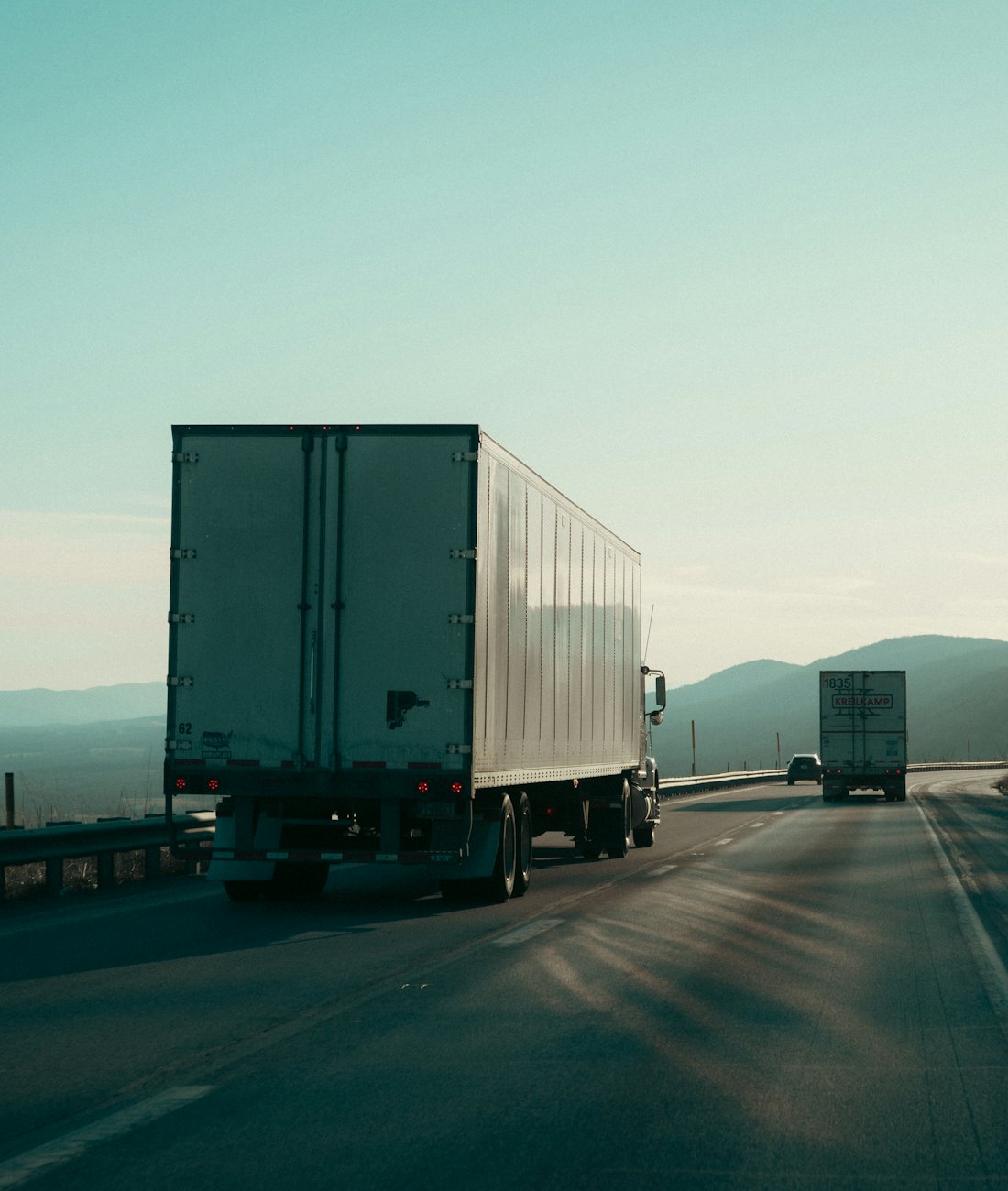 Two semi trucks with dry van trailers driving on a highway through the hills